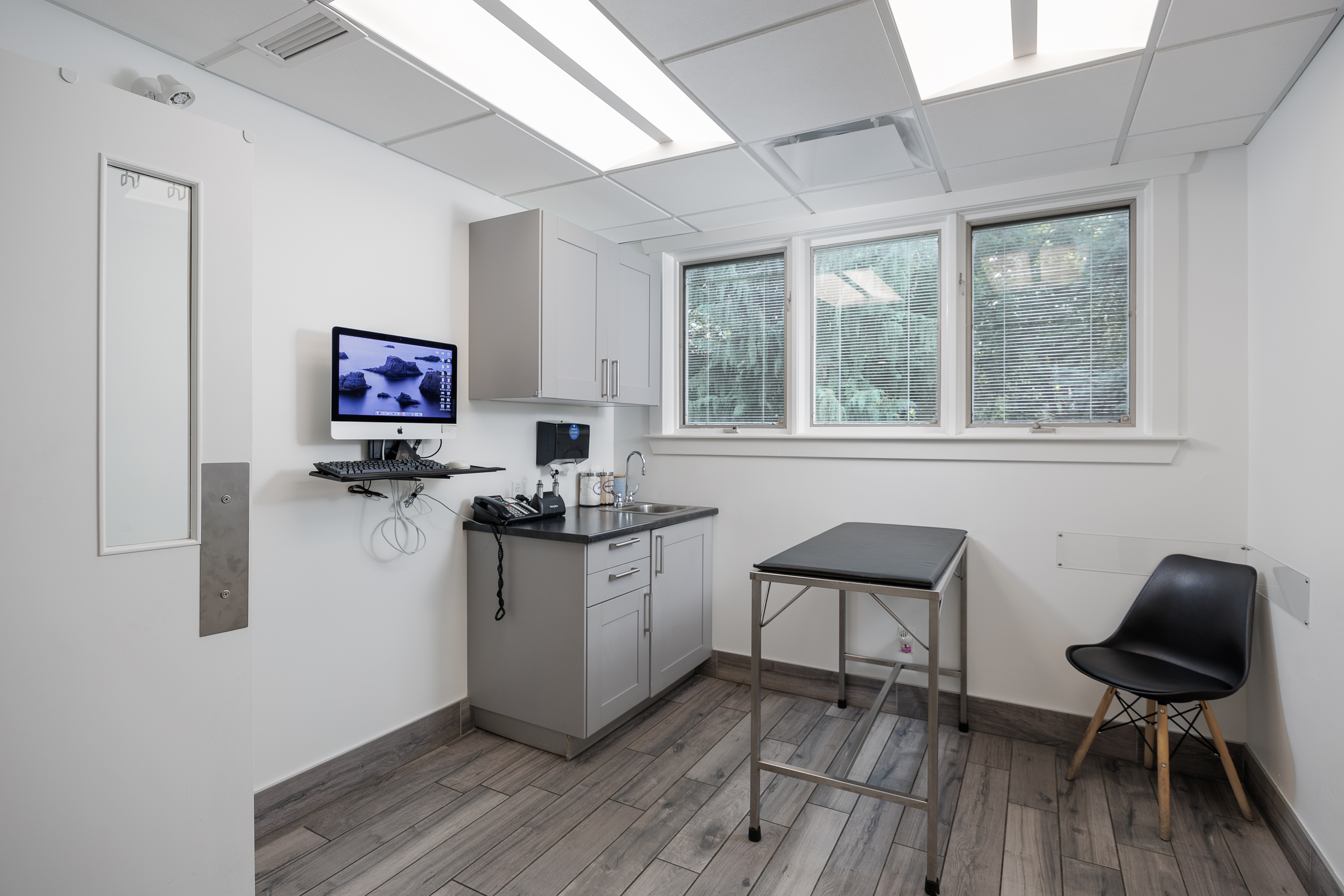 The image shows an interior view of a medical office with a modern design, featuring a clean room with white walls, a small desk area with a computer monitor, a chair, and a sink. There's also a window with natural light coming in.