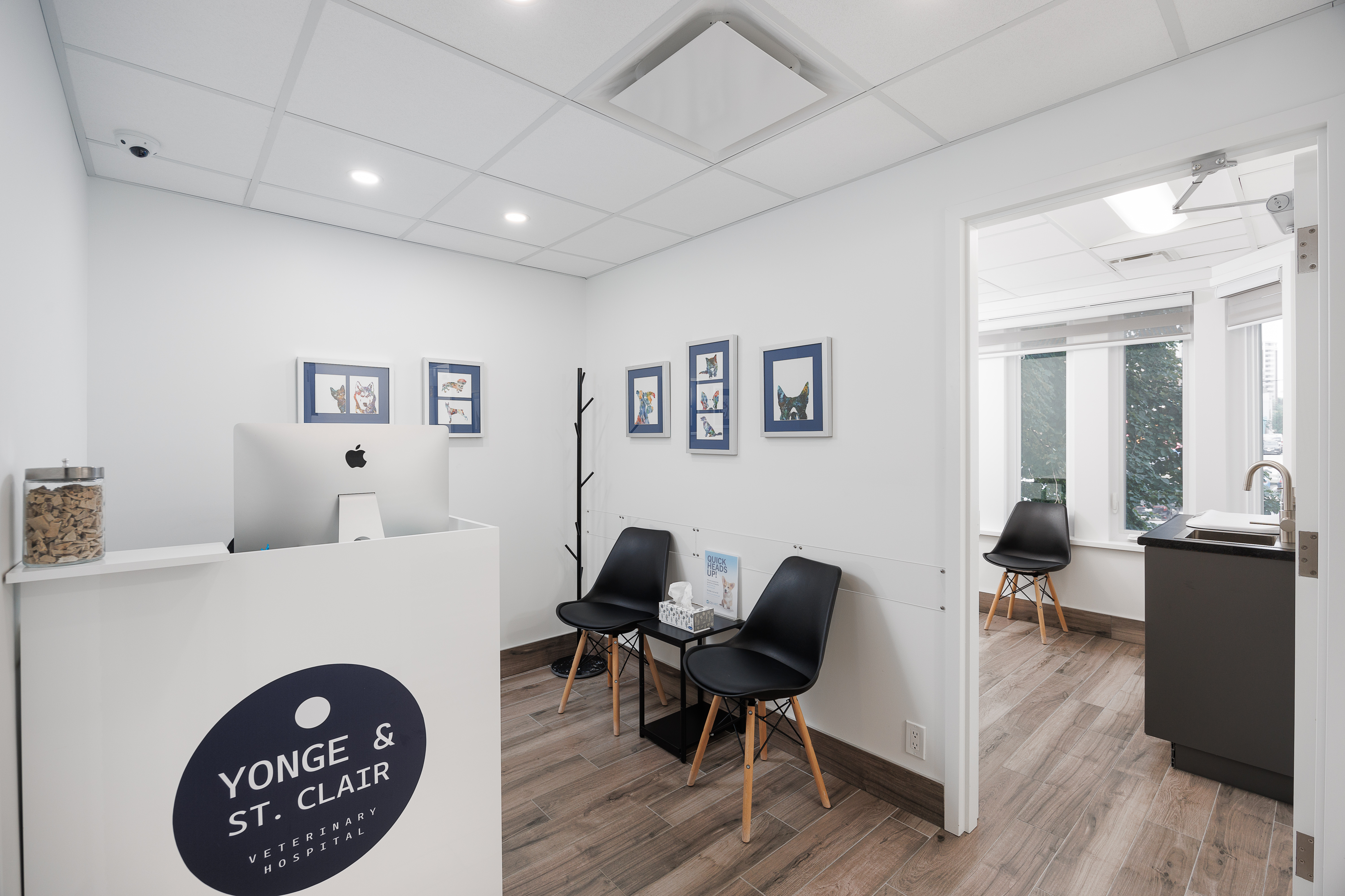 The image shows an interior view of a dental office with a clean and modern design, featuring a reception area with seating, a desk with a computer monitor and keyboard, a window with blinds partially drawn, and a sign that reads 'YONGE ST CLARE'.