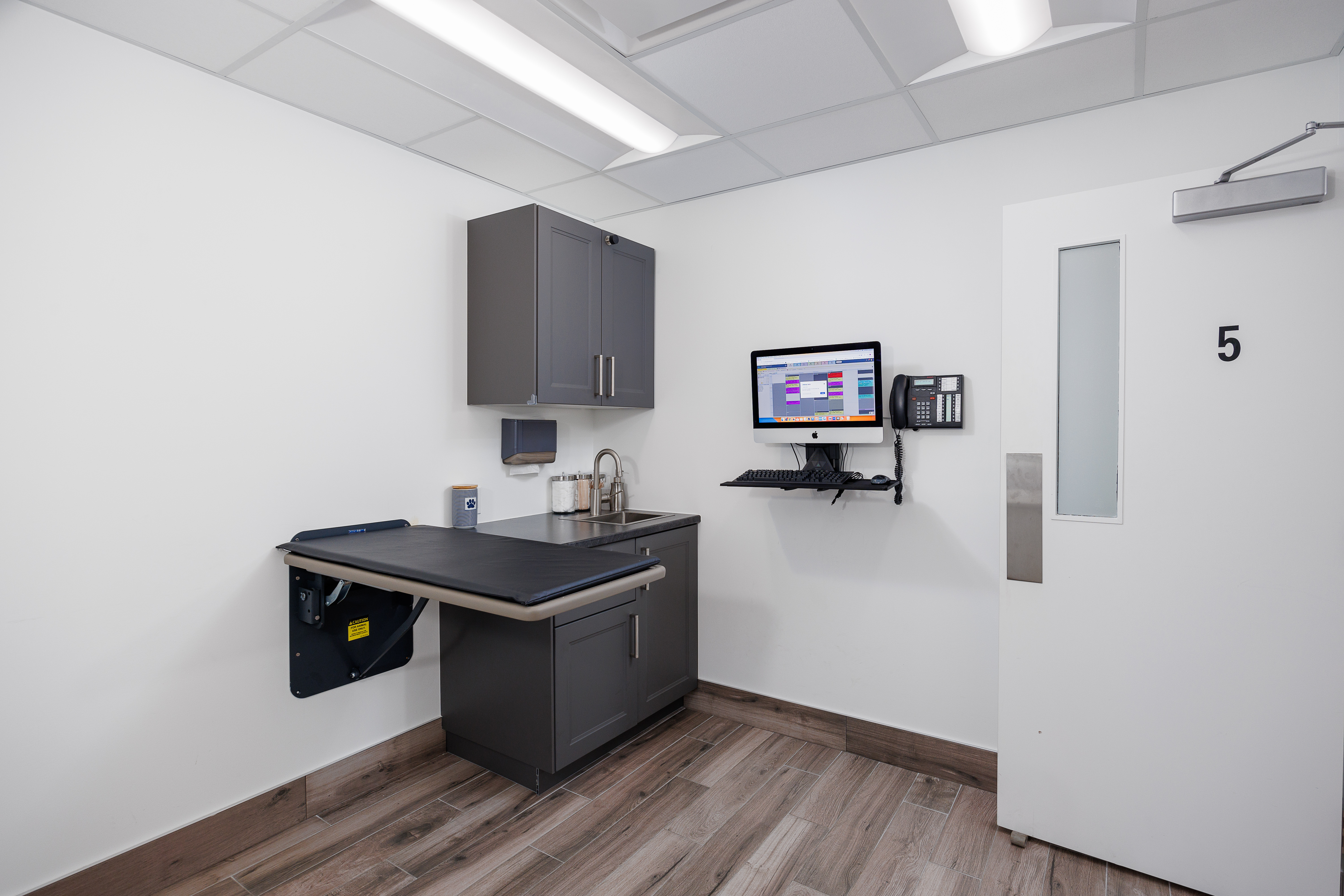 The image depicts a small, clean medical office with a desk holding a computer monitor, a sink, cabinets, and a chair.