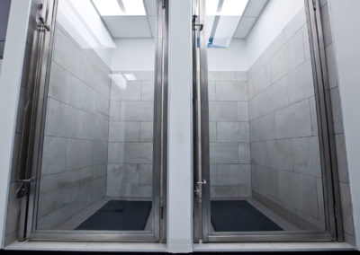 The image shows a modern, minimalist bathroom interior with white tiled walls, a gray floor, and stainless steel fixtures including a shower stall with clear glass doors.