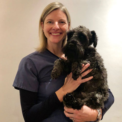 A woman wearing a blue shirt stands behind a black dog, both holding each other close.