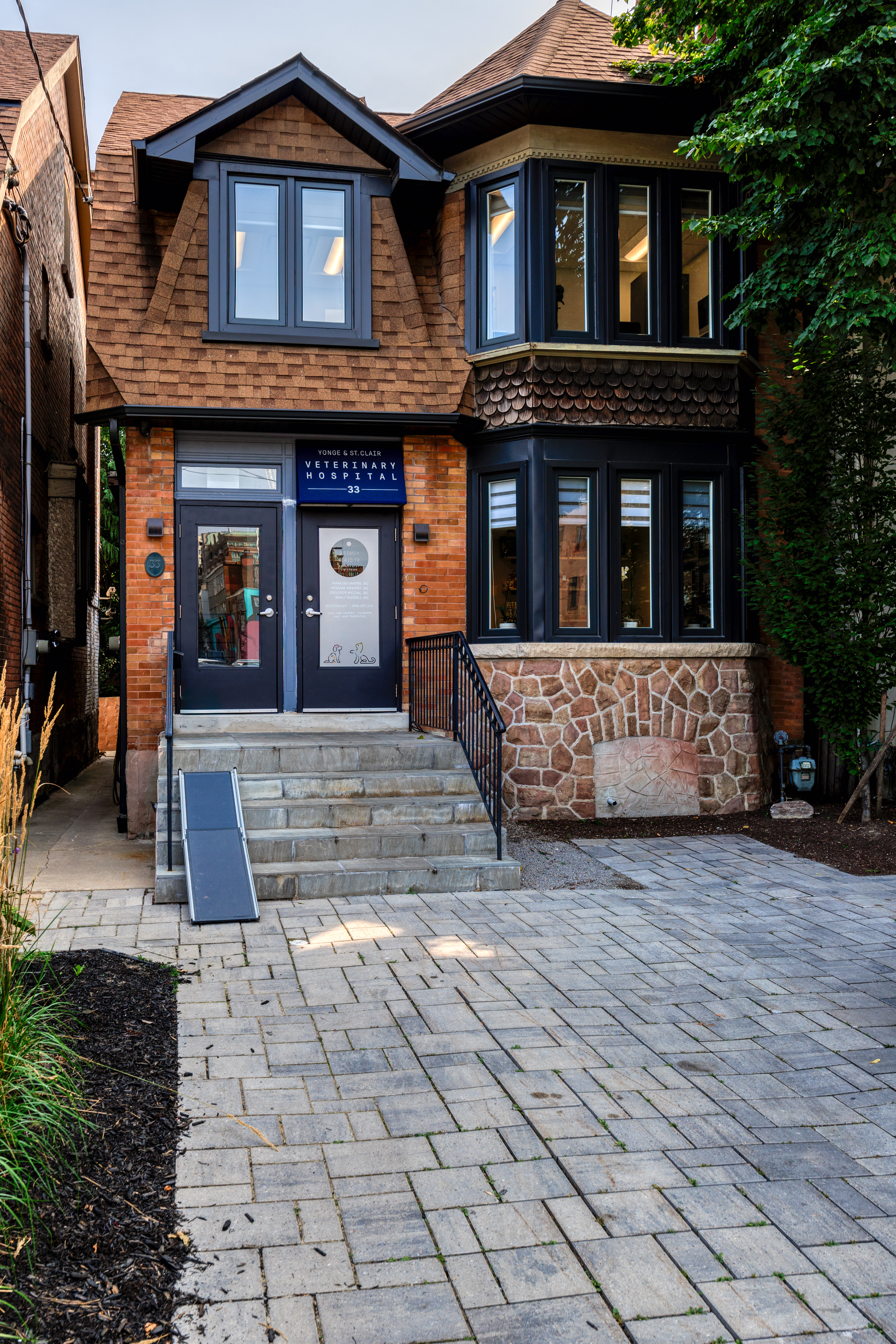 The image depicts a two-story residential building with a prominent front door, a small porch, and a brick pathway leading up to it. There are no visible people or vehicles in the scene.