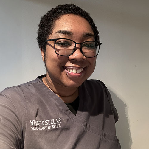 The image shows a smiling person wearing glasses, with short hair, posing for a photo against a plain background. They are dressed in a dark-colored scrub shirt that has text on it, indicating they may be a healthcare professional.