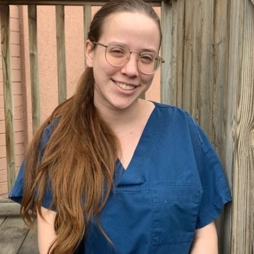 A woman wearing glasses, with her hair pulled back, stands smiling against a wooden railing.
